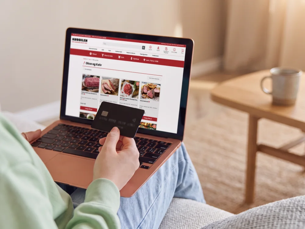 En person i sofaen med betalingskortet i hånden og Kødbilens webshop åben på den bærbare